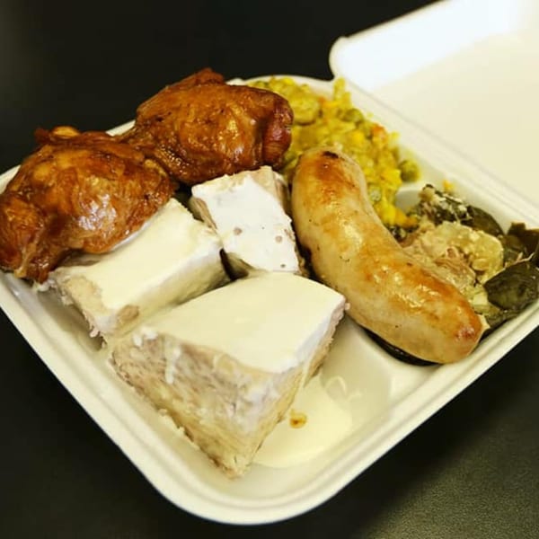 A Polynesian plate lunch with chicken, sausage, taro, and vegetables in a white takeout container.